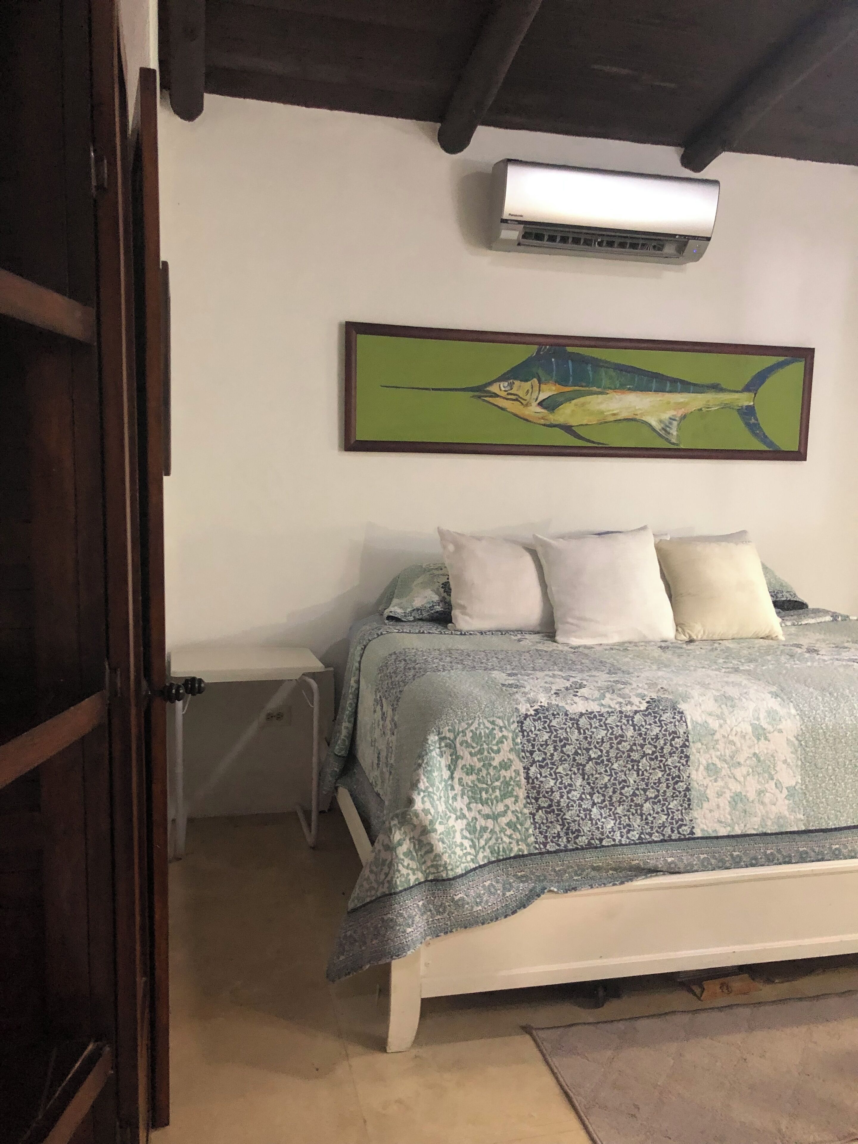 Een slaapkamer met een bed, kussens, een wandairconditioner en een lijst met een schilderij.