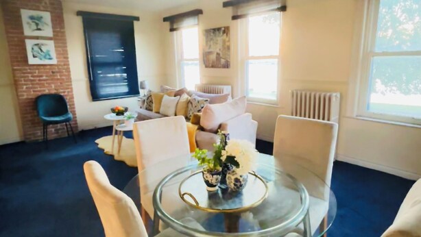 Un salon moderne avec une table en verre et des chaises blanches, un canapé avec des coussins, un mur en briques apparentes et des œuvres d’art.