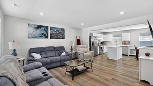 Un salon moderne avec un canapé gris, une table basse en verre et des œuvres d’art accrochées au mur. On aperçoit une cuisine à l’arrière-plan, avec des meubles blancs et un îlot central.