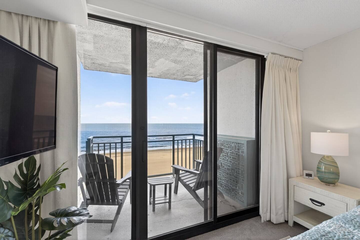 Un balcón con vista a la playa, una silla, una mesa, una lámpara y un televisor.