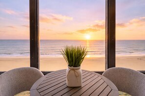 A view of the ocean at sunset through large windows, a round wooden table with a potted plant, and two chairs. - Beachfront Condo/ Pool/ Surfing/ Fishing (Virginia Beach)