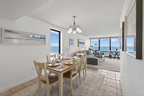 A spacious living area with a dining table set for four, a grey sofa, and a view of the ocean. - Beachfront Condo/ Pool/ Surfing/ Fishing (Virginia Beach)