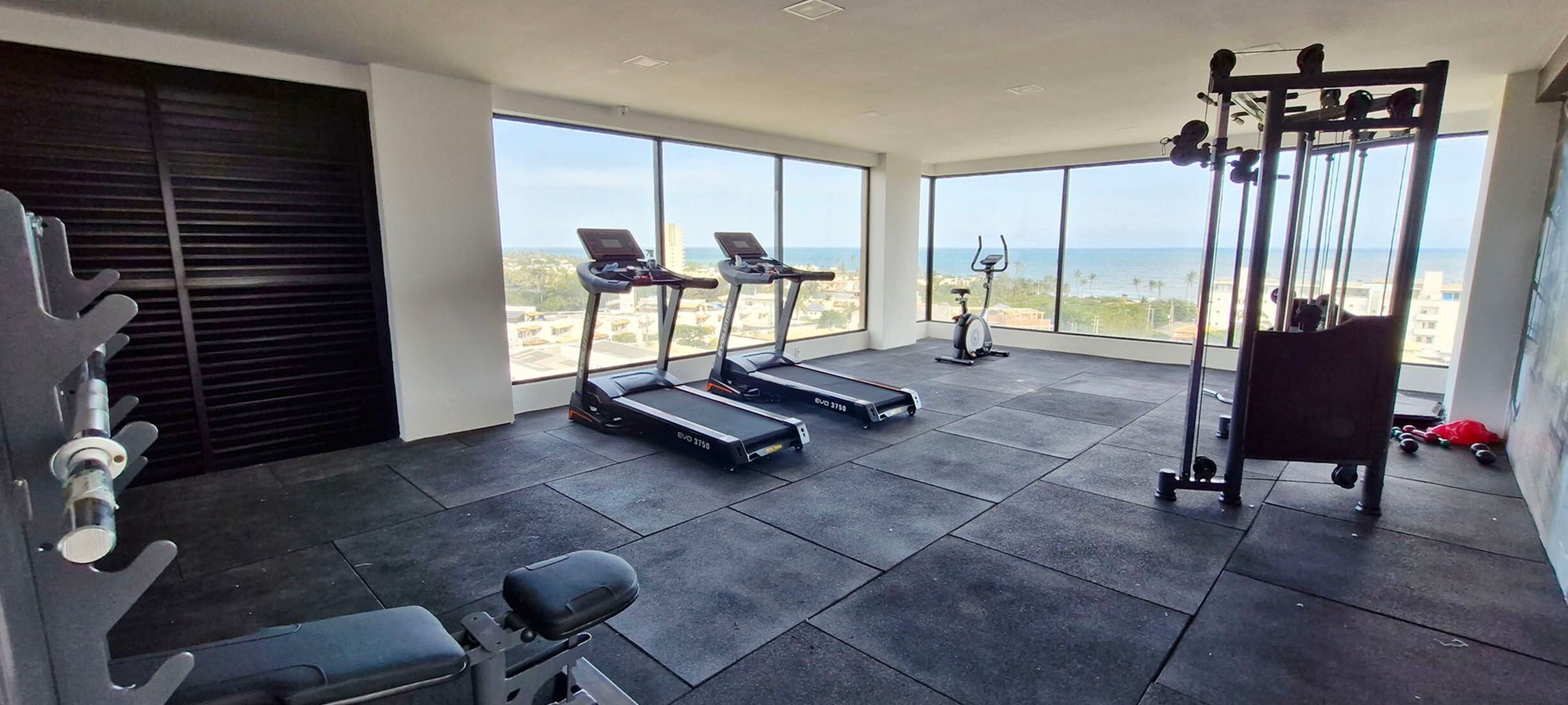 A gym with treadmills, an elliptical, and weightlifting equipment, large windows with a city view.