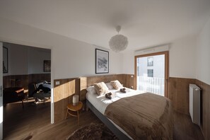 A modern bedroom with a large bed, wooden paneling, a small side table, a fluffy rug, and a balcony with a railing. - Three trips, one single stay on the gate of Paris ! You'll love it. (Bagneux)