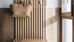 Een houten bank met een rieten mand er aan, voor een houten lattenwand en een raam.