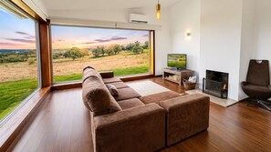 Ein modernes Wohnzimmer mit einer großen Eckcouch, einem Kamin, einem Fernseher und Blick auf ein Feld durch große Fenster.