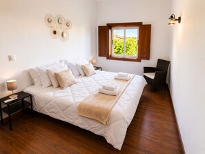 A bedroom with a large bed, a window with a view, a wall-mounted light fixture, and a small table with a lamp.