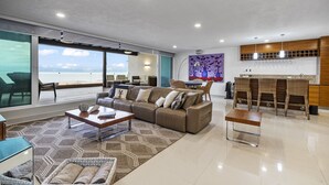 A spacious living area with a large sofa, a coffee table, and a dining area with wicker chairs. There is a large window offering a view of the ocean. - Resort-Style Villa w/ Private Padel & Infinity Pool (Bucerías)