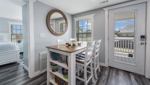 A dining area with a wooden table, white chairs, a woven mirror, and a view of the outside through a door. - 6 Seat Golf Cart~Family Friendly~Near DT Beaufort! (Beaufort)