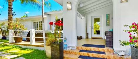 A white building with a covered porch, steps leading to the entrance, and a well-maintained garden with palm trees and potted plants.