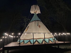 Une grande tente blanche en forme de pyramide, ornée d’une décoration suspendue, entourée d’une barrière métallique et illuminée de guirlandes lumineuses.