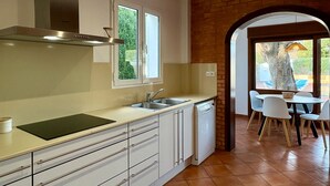 Een moderne keuken met witte kastjes, een roestvrijstalen afzuigkap, een raam en een vaatwasser. Door een boog is een eethoek zichtbaar met een tafel en stoelen.