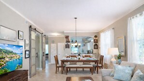 Un amplio salón con comedor, una mesa de centro con revistas, un sofá con cojines y un televisor que muestra un paisaje pintoresco.