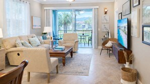 Un salón amplio con un sofá color beige, una mesa de centro y un televisor. Hay un balcón con vistas a palmeras y una montaña.