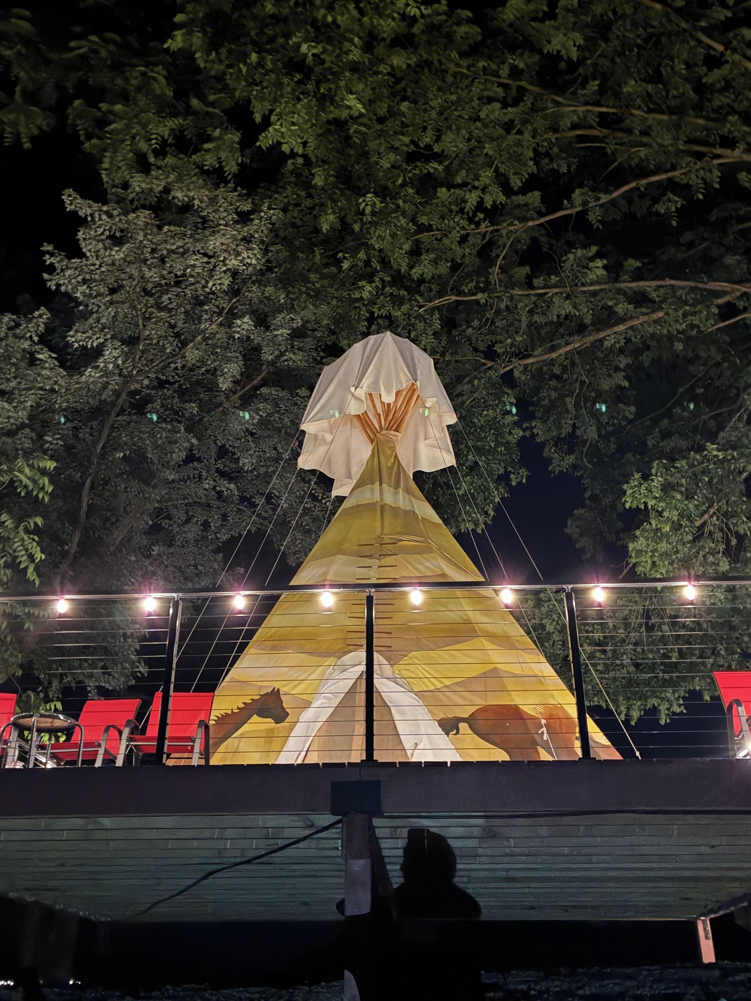 A large, illuminated tent structure with a pyramid-like shape, surrounded by trees and red seating areas.