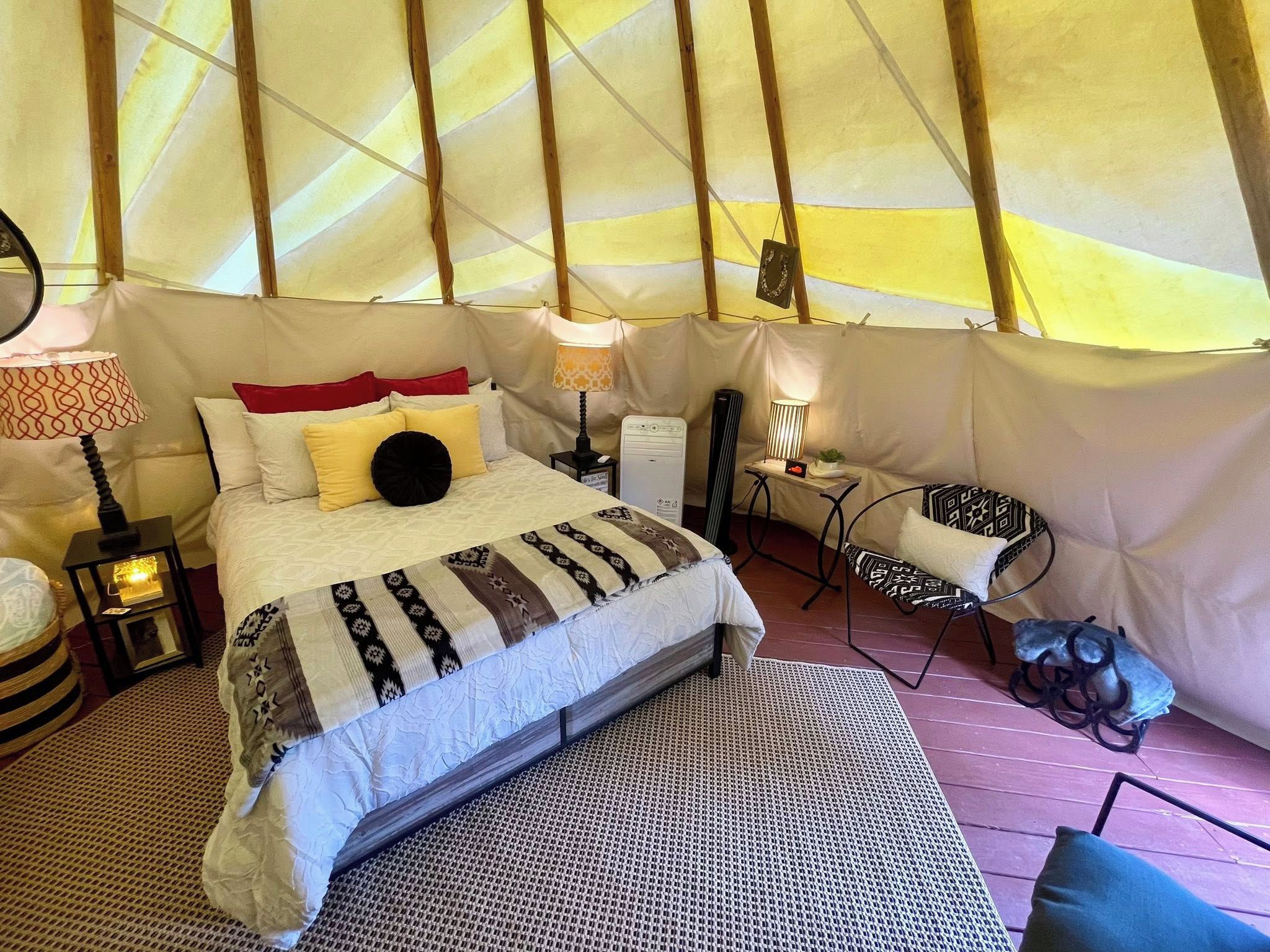A tented accommodation with a bed, bedside lamps, a small table, a chair, and a rug.