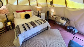 A tented accommodation with a bed, bedside lamps, a small table, a chair, and a rug.