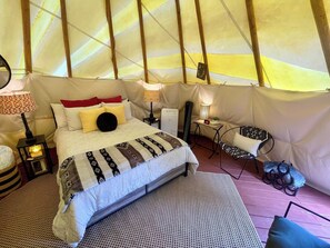 A tented accommodation with a bed, bedside lamps, a small table, a chair, and a rug.