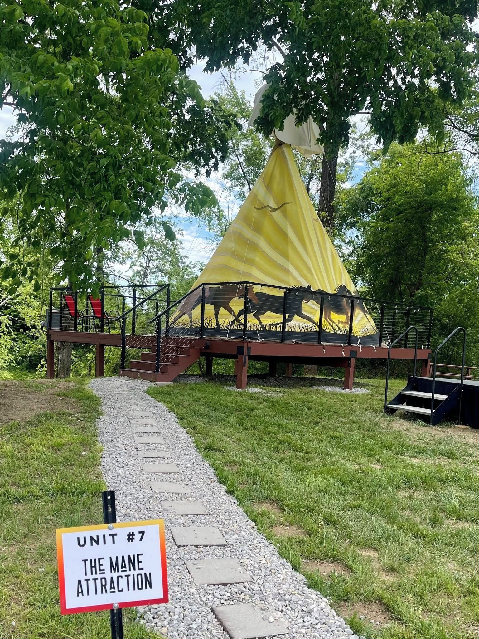 Une tente surmontée d’une auvent jaune, une pancarte indiquant « STAND N° 7, L’ATTRACTION PRINCIPALE », et un chemin de gravier menant à la tente.