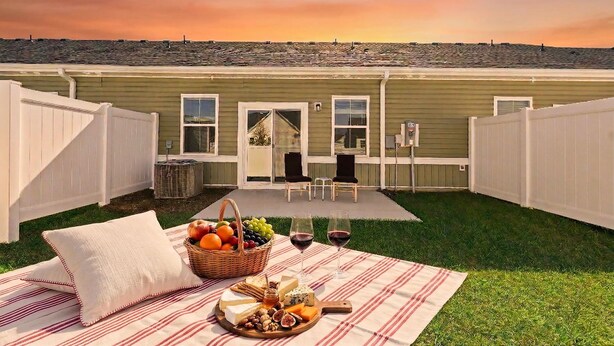 A backyard with a picnic setup, including a blanket, food, and drinks, and a view of a house with a deck and chairs. - New Modern 3BR/3BA Townhome. Quiet Retreat w/Pool, Playground and Free Parking. (Conway)