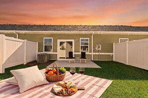 A backyard with a picnic setup, including a blanket, food, and drinks, and a view of a house with a deck and chairs.