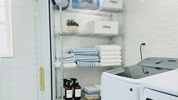 Un lavadero moderno con cabina de ducha de vidrio, lavadora blanca y estantes ordenados con toallas y cajas de almacenamiento.