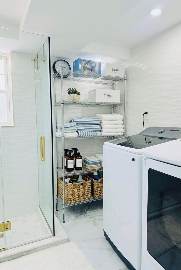A modern laundry room with a glass shower enclosure, a white washing machine, and neatly organized shelves with towels and storage boxes.