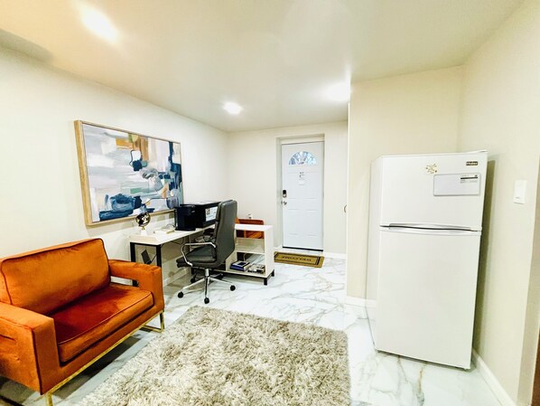 A compact living space with a sofa, a desk with a computer, a refrigerator, and a door.