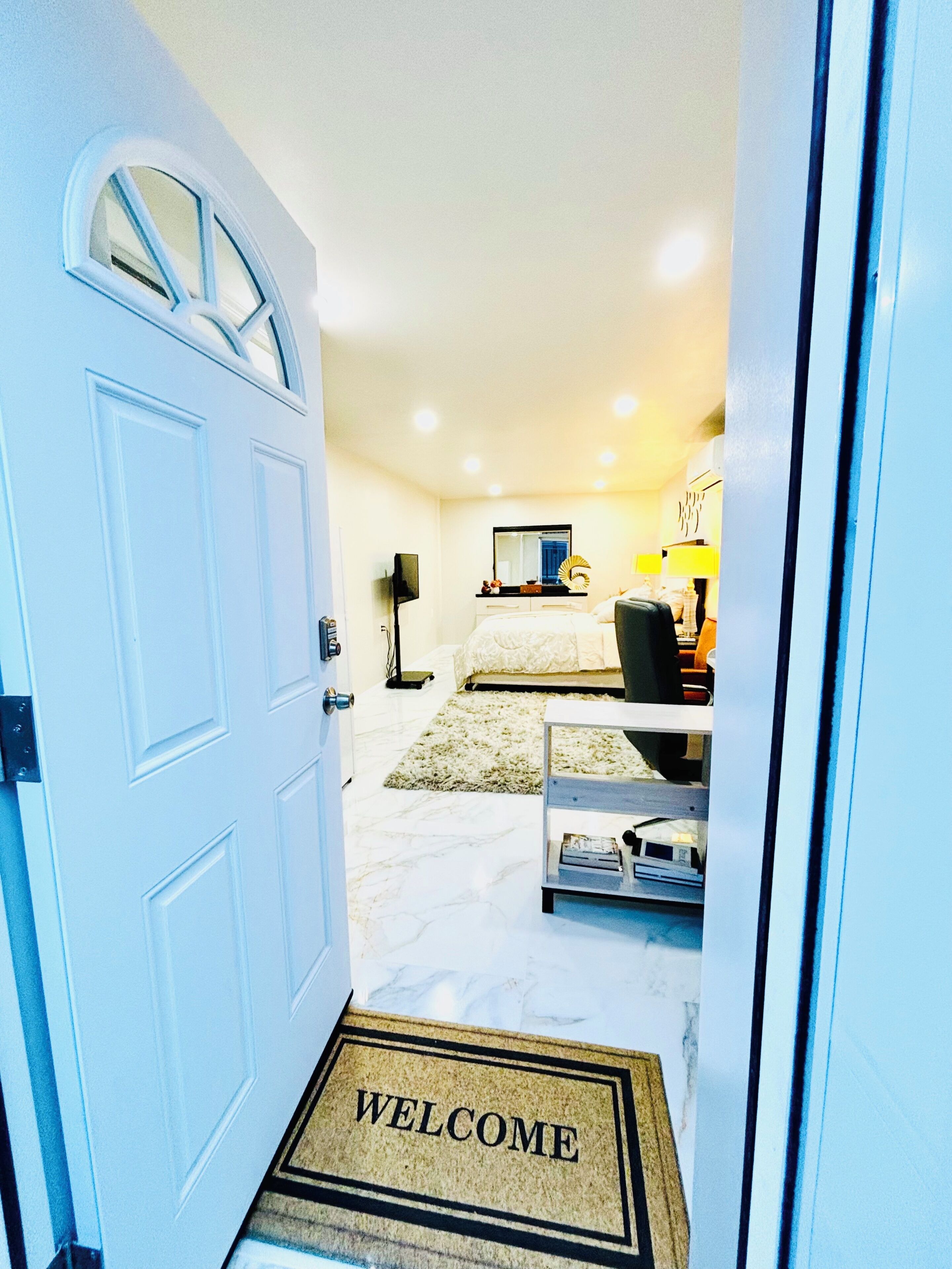 A hotel room with a white door, a welcome mat, a bed, a desk, a chair, a lamp, and a mirror.