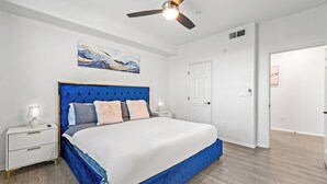 A bedroom with a large bed, a ceiling fan, a nightstand with a lamp, and a door. - Modern 2BR Apt + Mins to Disney + Arcade (Placentia)