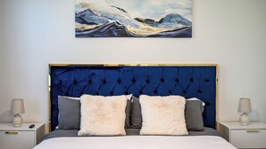 A bedroom with a large bed, a blue upholstered headboard, and two bedside tables with lamps. - Modern 2BR Apt + Mins to Disney + Arcade (Placentia)