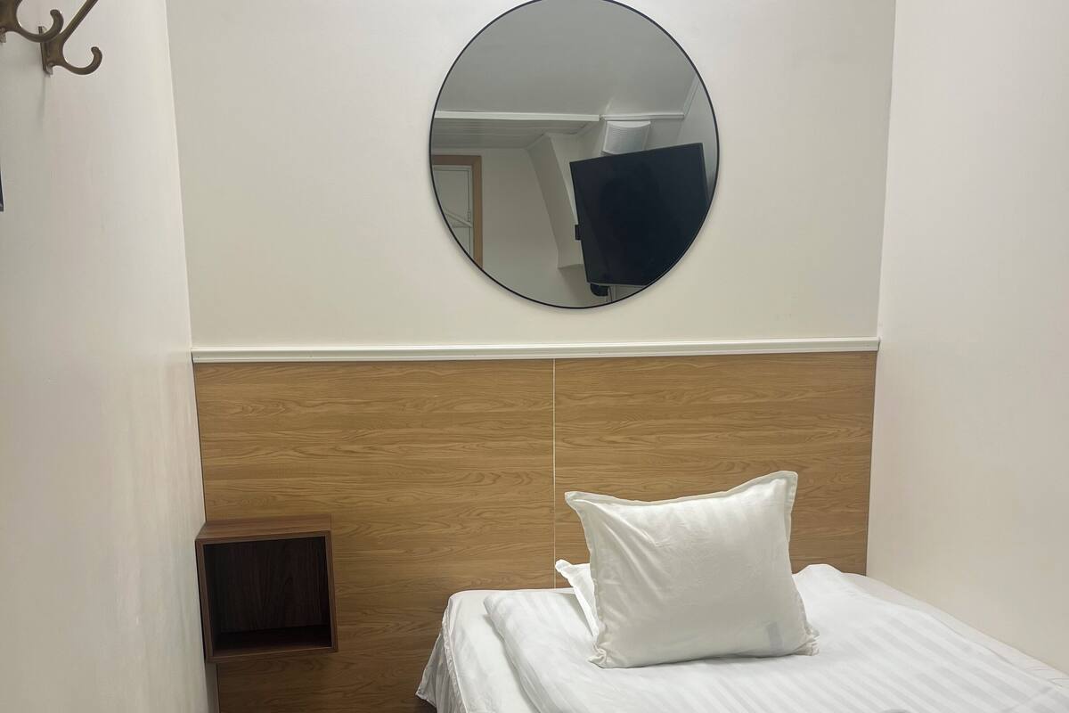 Ein Hotelzimmer mit einem Holzkopfteil, einem runden Spiegel, einem Bett mit weißer Bettwäsche und einer kleinen Holzkabine.