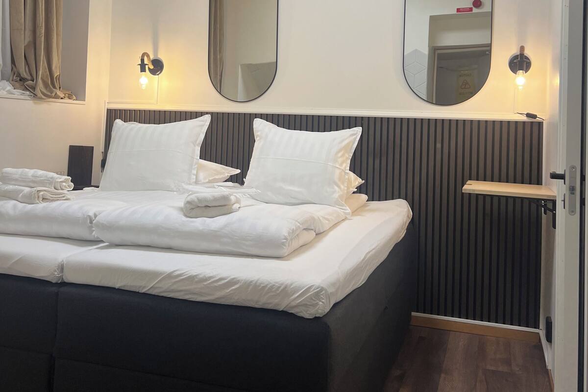 Ein ordentlich bezogenes Bett mit weißen Leinentüchern, zwei an der Wand befestigte Spiegel und ein Nachttisch mit Lampe.