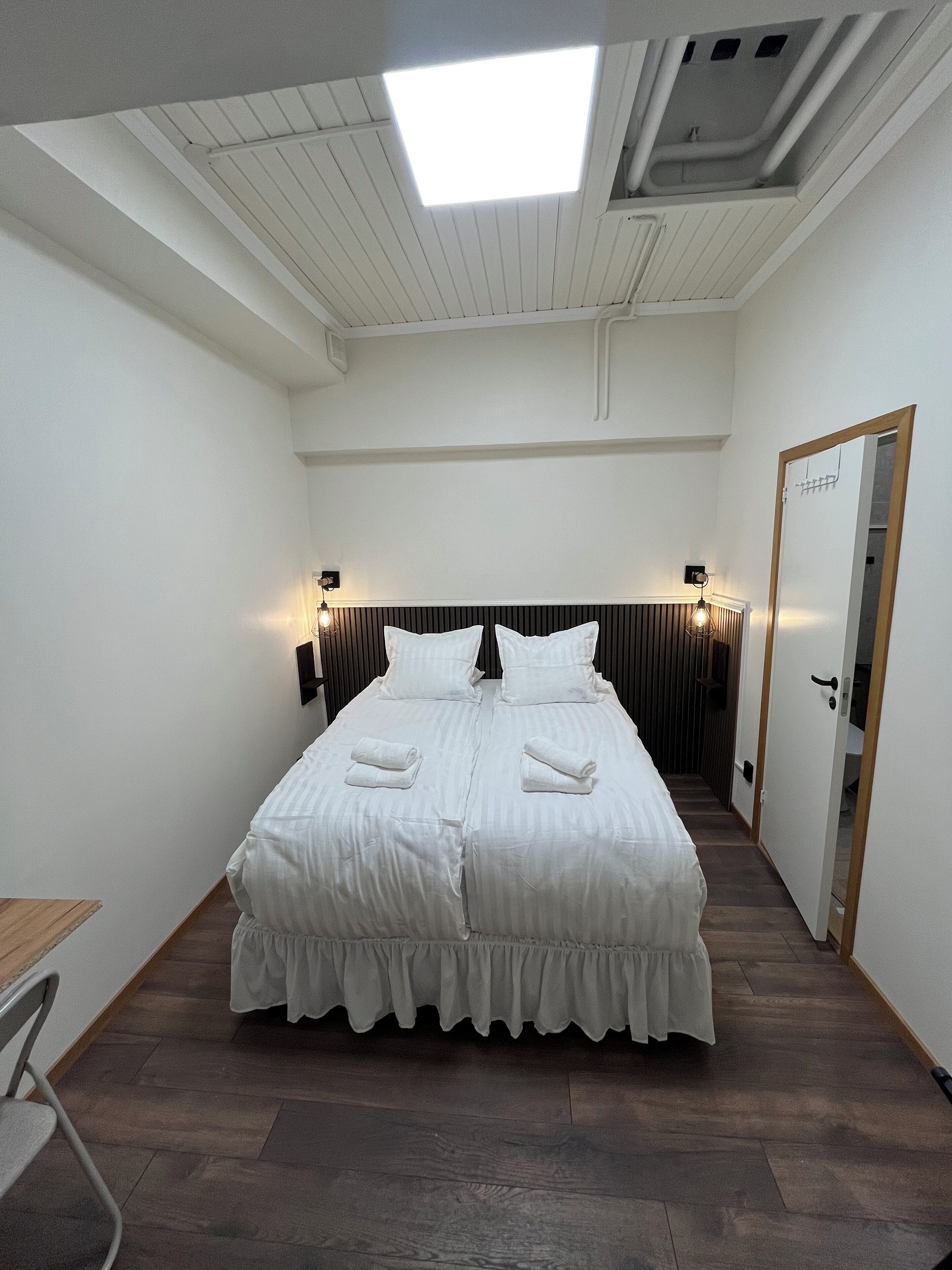 Una habitación de hotel con una cama grande, dos mesitas de noche, una lámpara en el techo y una puerta.