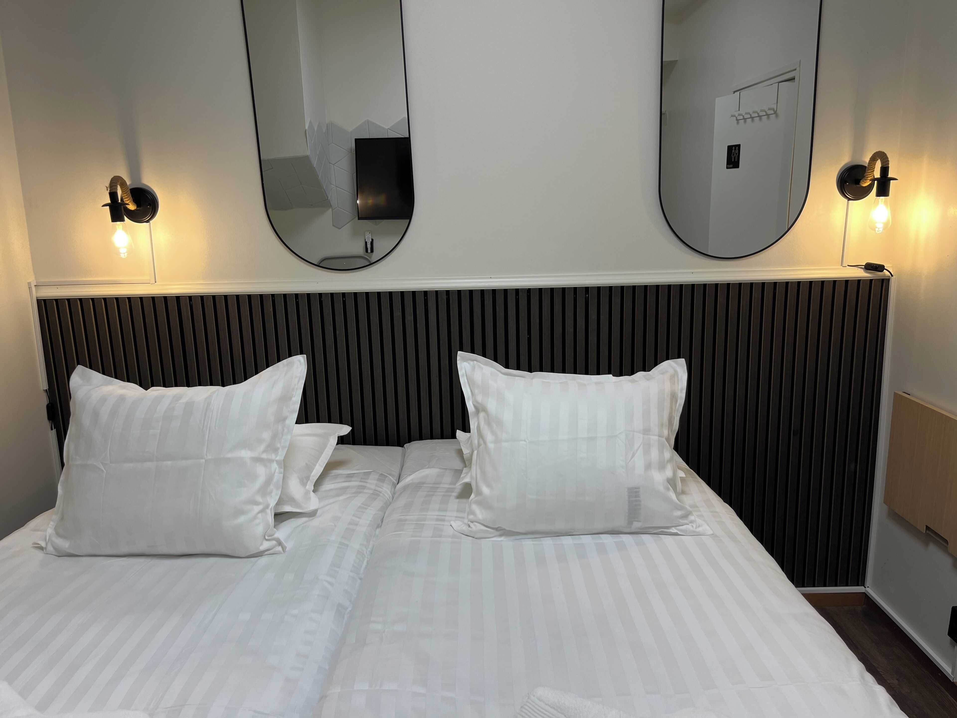 Una habitación de hotel con una cama grande, dos mesitas de noche, un televisor montado en la pared y dos apliques.