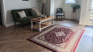 Una sala de estar con un sofá verde, una mesa de centro de madera, una alfombra estampada, una lámpara, una silla y una planta en maceta.
