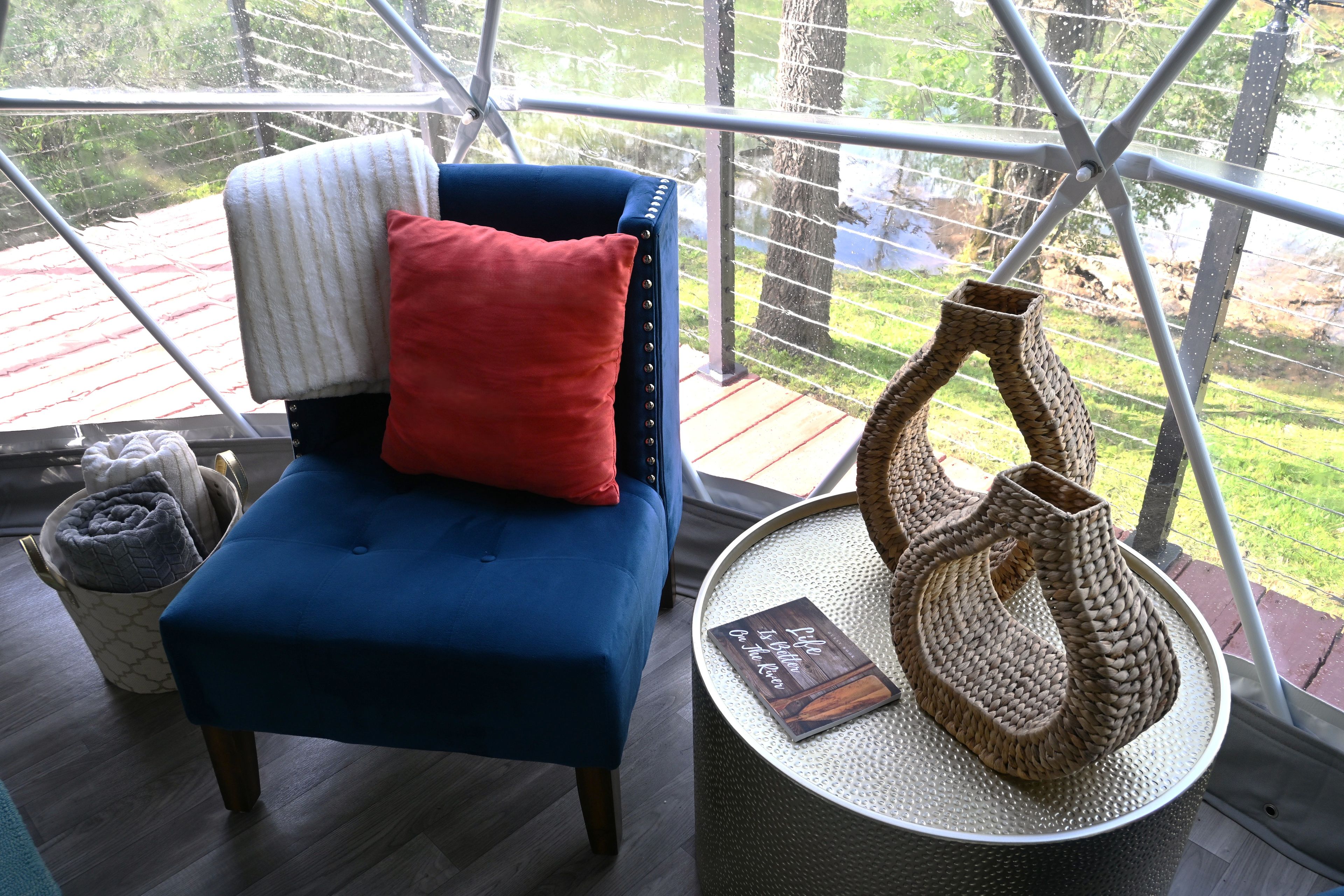 Una silla azul con un cojín rojo, una cesta de mimbre y un libro sobre una mesa redonda.