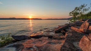A sunset over a calm lake with large rocks in the foreground and a distant mountain range. - Naut on Call Geodome at The Edge—Unique Riverfront Glamping in Salt Lick, KY (Salt Lick)