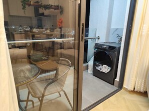 Ein Balkon mit Glastür, einem runden Tisch mit Stühlen, einer Waschmaschine und Blick auf die Stadt.