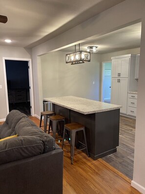 A modern kitchen with a bar area, wooden flooring, and a gray sofa in the living area. - Spacious 4-Bedroom Modern Farmhouse Ideal Family Getaway & Gatherings (Rolla)