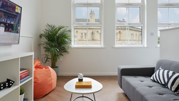 Een moderne woonkamer met een grijze bank, een ronde witte salontafel en een groot raam met uitzicht op gebouwen.