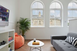 Ein modernes Wohnzimmer mit einem grauen Sofa, einem runden weißen Couchtisch und einem großen Fenster mit Blick auf Gebäude.