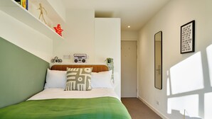 Een slaapkamer met een bed, een plank met boeken en een radio, een spiegel en een lijst met een citaat aan de muur.