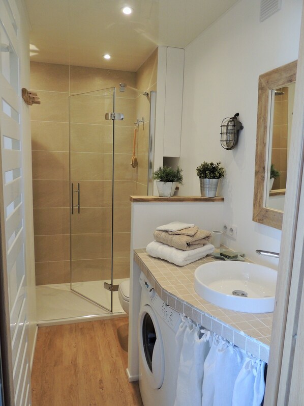 A modern bathroom with a glass shower enclosure, a built-in washing machine, a sink, and a mirror.