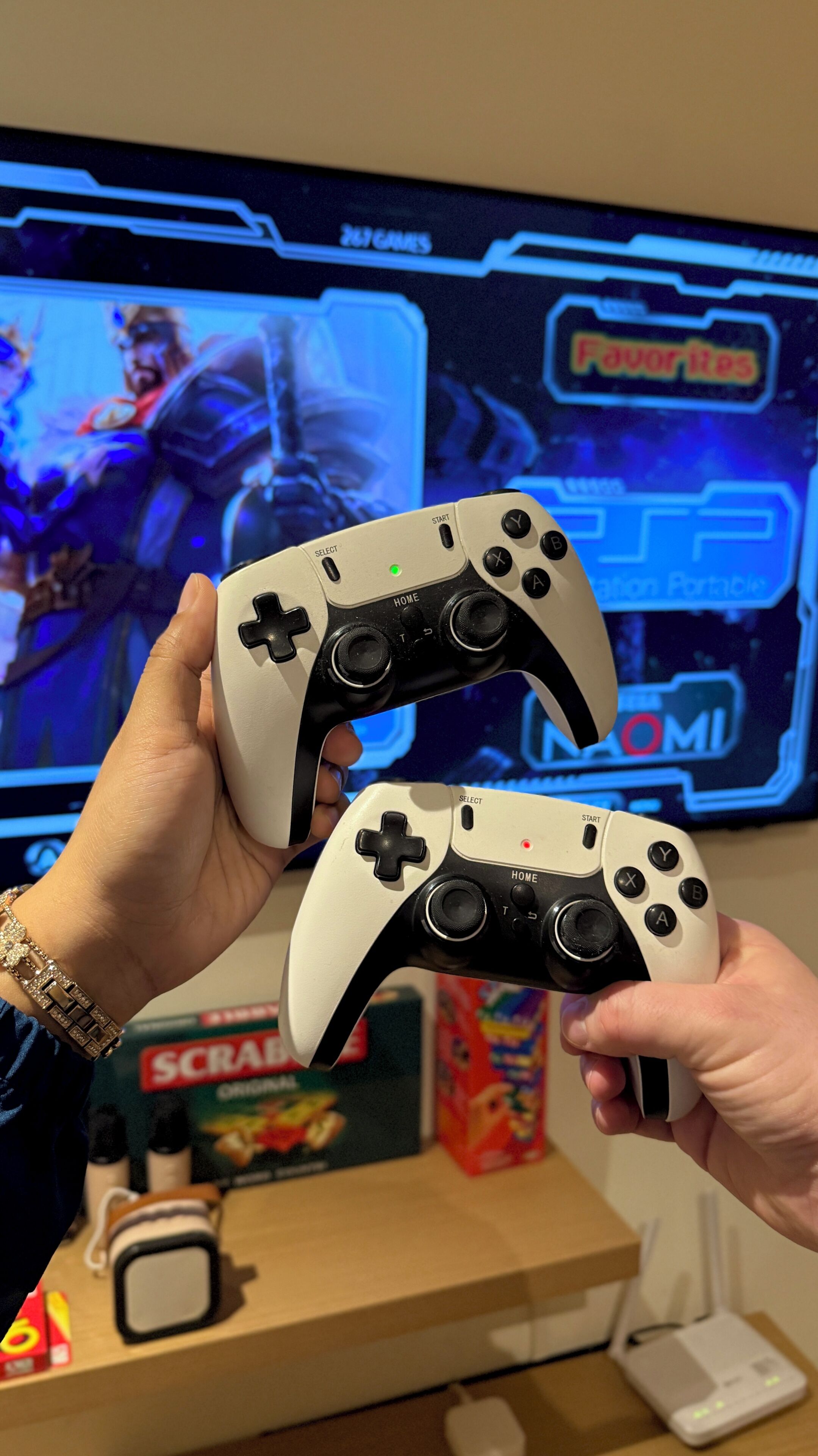 Two game controllers held by a person. In the background, there is a television displaying a video game, a box labeled "SCRABBLE," and a white router on a table.