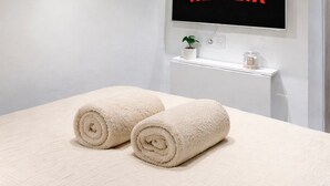 Moderni hotellihuone, jossa on sänky, kaksi rullalle taitettua pyyhettä, seinälle asennettu televisio, jossa näkyy teksti »NETFLIX», pieni kasvi ja valkoinen hylly.