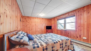 A bedroom with wooden walls, a bed with a checkered bedspread, a wooden nightstand, a television, and a window with a view of the outside.