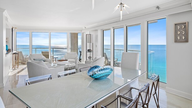Una habitación amplia con una gran mesa de vidrio, muebles blancos y vista al océano.