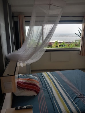 A room with a bed, a window with a view of the sea, and a ceiling fan. - Functional duplex with superb sea view (Les Trois-Îlets)