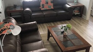 A living room with a brown leather sofa, a wooden coffee table, and a lamp. There are two windows with sheer curtains and a hammock hanging between them. - Quaint 2-bedroom house wanting your TLC in charming Hammondsport (Hammondsport)